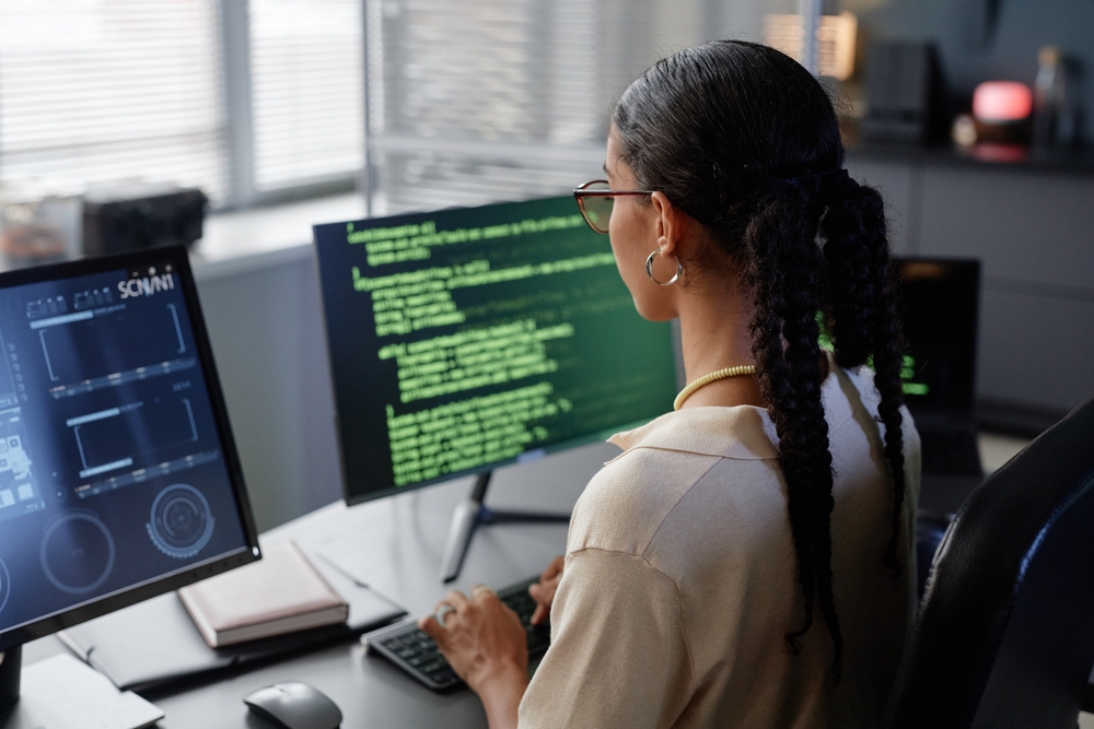 Rear view of female programmer with glasses reading multiple computer screens while working on code at professional workstation in IT company office